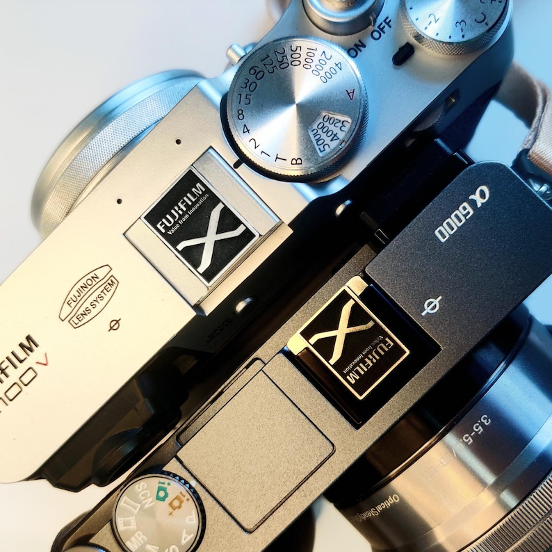 May include: A silver and black digital camera with a silver lens and a black lens cap. The camera has a black strap and a black and silver flash. The camera has a black and silver dial with the text "FUJIFILM" and "X-G000" on it.
