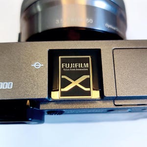 May include: A close-up of a silver camera with a black lens and a gold Fujifilm X logo on the top. The camera has a black and gold inscription that reads "FUJIFILM Value from Innovation".