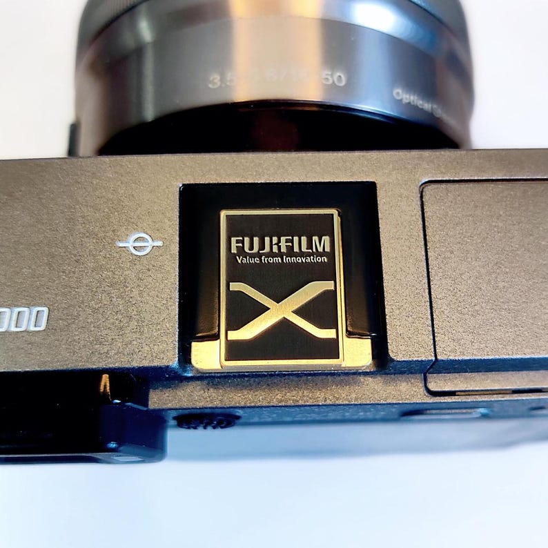 May include: A close-up of a silver camera with a black lens and a gold Fujifilm X logo on the top. The camera has a black and gold inscription that reads "FUJIFILM Value from Innovation".