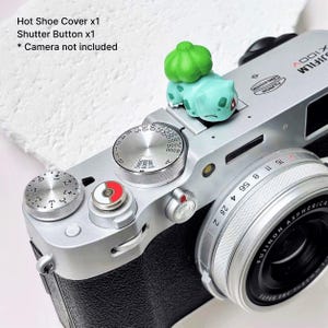May include: A silver Fujifilm X100V camera with a green and blue Bulbasaur hot shoe cover and a red and white shutter button. The camera has a black lens and various dials and buttons. The text "Hot Shoe Cover x1, Shutter Button x1" is visible.