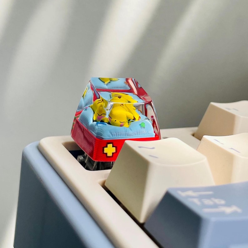 May include: A custom keyboard keycap featuring a yellow cartoon character resting on a blue and red bed. The keycap has a clear, transparent cover and a red cross symbol on the side. The keycap is on a light blue keyboard.