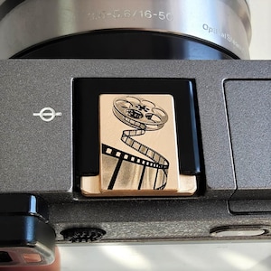 以下が含まれることがあります： グレーのカメラのクローズアップ。金色のフィルムリールデザイン。デザインはフィルムリールとフィルムストリップが特徴です。カメラレンズには「3.5-5.6/16-50」と「Optical SteadyShot」の文字があります。