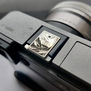 May include: A silver camera with a black and white engraved metal mountain scene hot shoe cover. The camera lens has the text "3.5-5.6/16-50" printed on it.