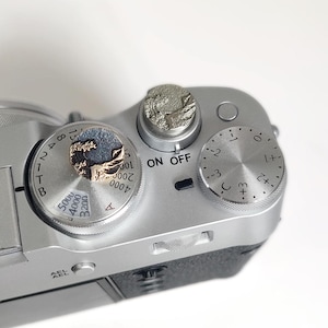 May include: A silver-colored camera top plate featuring two circular metal shutter release buttons with embossed wave designs. One button is gold-toned with a dark wave, while the other is silver-toned. The camera has dials with numbers and the text ON OFF.