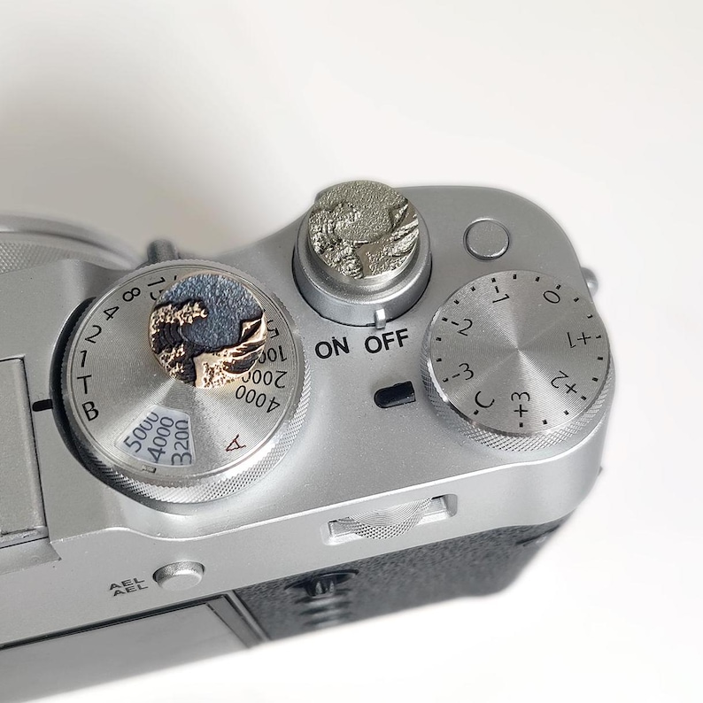 May include: A silver-colored camera top plate featuring two circular metal shutter release buttons with embossed wave designs. One button is gold-toned with a dark wave, while the other is silver-toned. The camera has dials with numbers and the text ON OFF.