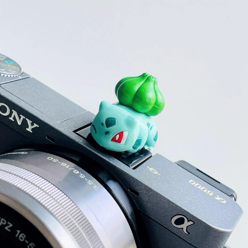 May include: A light blue Bulbasaur figurine with a green bulb on its back, perched on a gray Sony camera. The figurine has red accents on its face and is a collectible item.