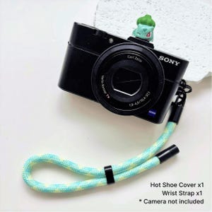 May include: Black Sony digital camera with a Carl Zeiss lens and a green and blue wrist strap. A small green figurine sits on top of the camera. The image includes text that reads "Hot Shoe Cover x1, Wrist Strap x1, * Camera not included."
