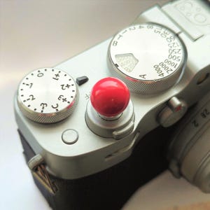 May include: A silver digital camera top plate featuring a bright red circular shutter release button, a silver exposure compensation dial with numerical markings from -3 to +3, and a silver shutter speed dial with settings from 1 to 4000.