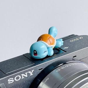 Puede incluir: Una pequeña figura de tortuga azul claro con un caparazón color canela y una expresión de guiño, colocada sobre el cuerpo de una cámara Sony Alpha 6000 gris oscuro. El objetivo plateado muestra el texto 3.5-5.6/16-50 y la marca Sony.