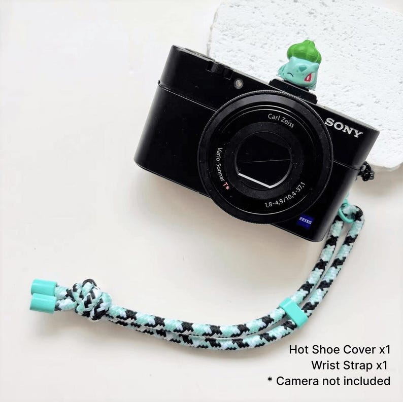 May include: A black Sony digital camera with a Carl Zeiss lens and a green and white patterned wrist strap. A small green figurine sits on top of the camera. The text on the image reads "Hot Shoe Cover x1, Wrist Strap x1, * Camera not included."
