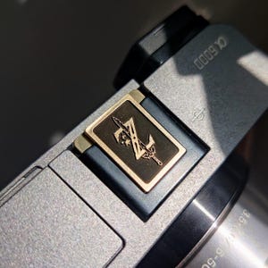 Puede incluir: Una cámara plateada con una placa de metal dorado en la parte superior. La placa tiene un fondo negro con una espada dorada y la letra "Z" en ella. La cámara tiene el texto "α6000" en la parte superior.