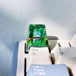 以下が含まれることがあります： 緑と青の漫画のキャラクターが描かれた、詳細なミニチュアシーンのカスタムキーキャップ。 透明なキーキャップは、赤いキノコと緑の葉のある風景の中にキャラクターを表示します。 白いキーボードに取り付けられています。