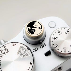 May include: Close-up of a silver camera with a black and white cat Yin Yang design on the on/off dial. The camera has multiple dials with numbers and letters. The on/off dial is gold-rimmed.