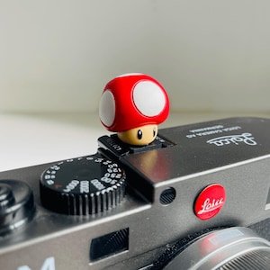 Puede incluir: Una figura de hongo rojo y blanco con una cara sonriente se encuentra sobre una cámara Leica. La cámara es gris con diales negros y el logotipo de Leica en rojo. La figura es un personaje popular de videojuegos.