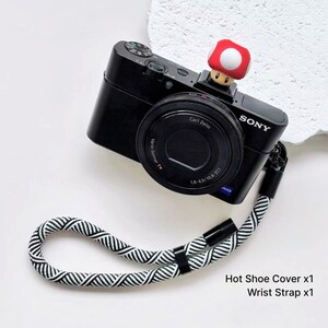 May include: A black Sony digital camera featuring a red and white mushroom-shaped hot shoe cover and a black and white braided rope wrist strap. The text Hot Shoe Cover x1 and Wrist Strap x1 is printed in the lower right corner of the image.