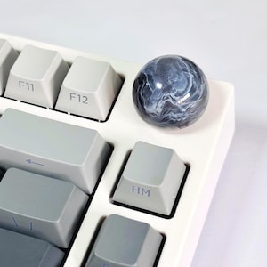 Puede incluir: Un teclado blanco con teclas grises y una esfera de mármol blanco y negro en la tecla 'HM'.