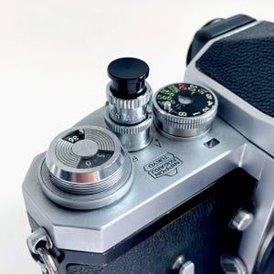 以下が含まれることがあります： 黒とシルバーのダイヤルと黒いトップを備えた、ヴィンテージのシルバーカメラのクローズアップ。 カメラはテクスチャ加工された黒いグリップを備え、「Nippon Kogaku Tokyo」の文字が見えます。