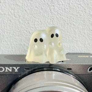 May include: A translucent, off-white resin figurine featuring two conjoined ghost shapes with simple black circular eyes. The small, smooth object is positioned on the top surface of a black camera body, resting near the lens mount area.