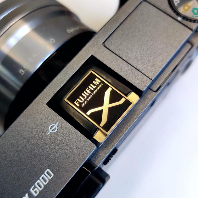 May include: A close-up of a gray camera with a gold "FUJIFILM Value from Innovation" logo and a black "X" logo.
