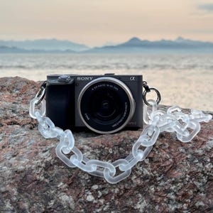May include: A dark gray Sony Alpha camera with a clear, chain-link style strap. The camera lens is centered, and the strap is draped across a rock. The background features a body of water and a hazy sky.