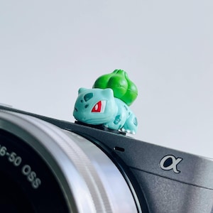 May include: A small, light blue Bulbasaur figurine with red eyes and a green bulb on its back rests on a gray camera. The camera has a silver lens and the brand logo "&alpha;" is visible.