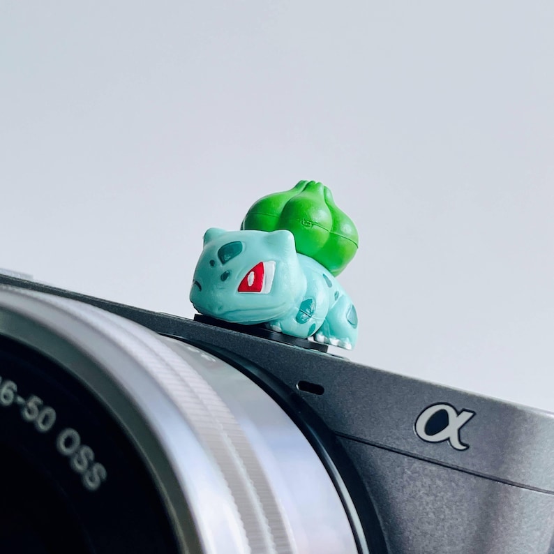 May include: A small, light blue Bulbasaur figurine with red eyes and a green bulb on its back rests on a gray camera. The camera has a silver lens and the brand logo "&alpha;" is visible.