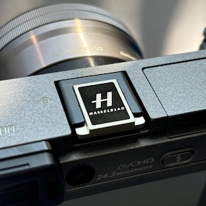 May include: A close-up of a silver camera with a black Hasselblad logo on the side. The camera has a lens cap on and the text "24.3 Megapixels" and "AVCHD" are visible.