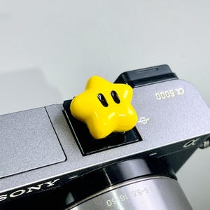 May include: A bright yellow, star-shaped hot shoe cover with two black oval eyes, attached to the top of a grey Sony Alpha 6000 camera. The camera body features the text Sony and Alpha 6000, with a lens showing 3.5-5.6/16-50 specifications.