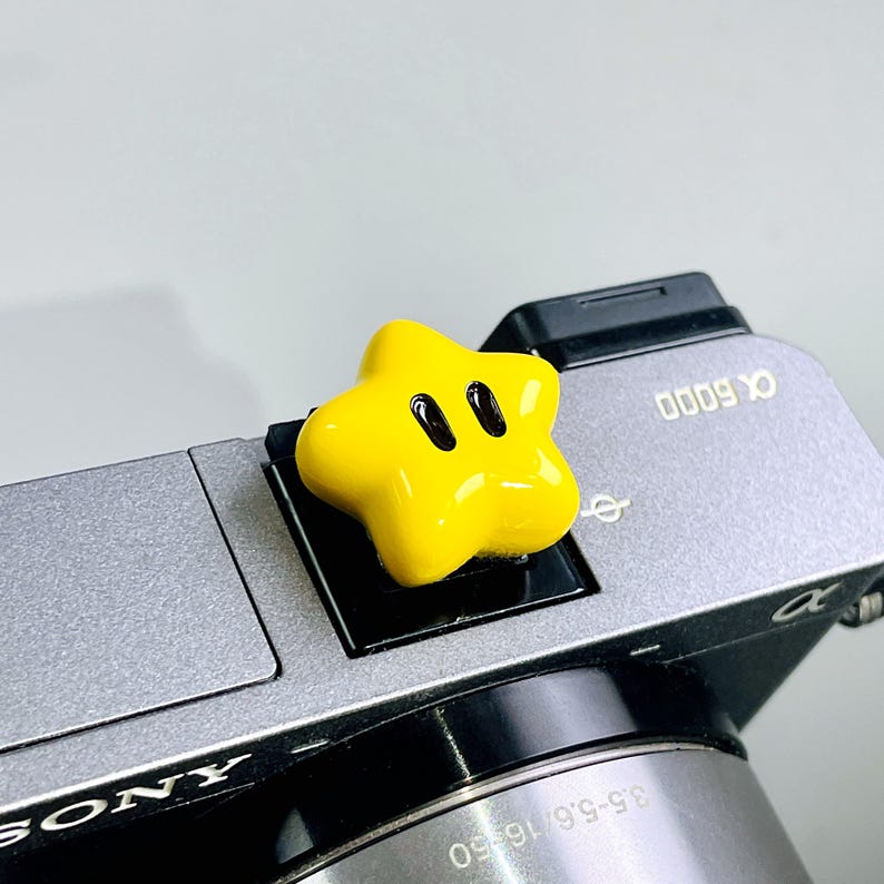May include: A bright yellow, star-shaped hot shoe cover with two black oval eyes, attached to the top of a grey Sony Alpha 6000 camera. The camera body features the text Sony and Alpha 6000, with a lens showing 3.5-5.6/16-50 specifications.