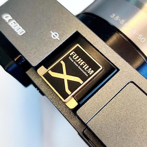 May include: A close-up of a silver camera with a black lens and a gold Fujifilm X logo. The camera has a black and gold Fujifilm X logo on the side. The text "Value from Innovation" is printed below the logo.