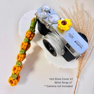 May include: A silver and black Fujifilm X100V camera featuring a yellow flower-shaped hot shoe cover and a long, crocheted wrist strap made of orange and green yarn. The strap consists of a repeating pattern of orange flowers with green centers.