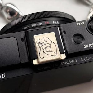 Puede incluir: Una cámara digital negra con una lente y una cubierta decorativa para la zapata del flash. La cubierta es dorada y presenta un dibujo lineal de una mujer abrazando una luna creciente. La lente tiene el texto "Vario-Sonnar T* F1.8".