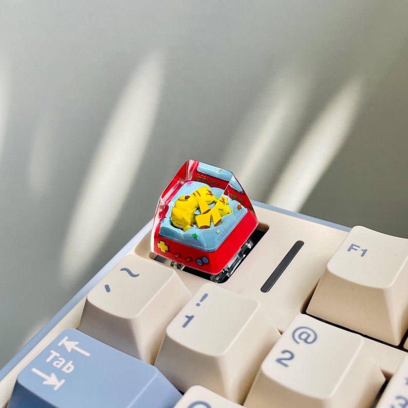 May include: A custom keyboard keycap featuring a miniature, colorful video game console design. The keycap is red, blue, and yellow, with a clear resin shell. It is installed on a light-colored keyboard.