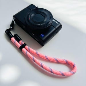 Puede incluir: Una cámara digital Sony negra con lente Zeiss y una correa de muñeca de cuerda rosa y morada. La cámara está sobre una superficie blanca. La correa de muñeca está hecha de una cuerda rosa con rayas moradas.