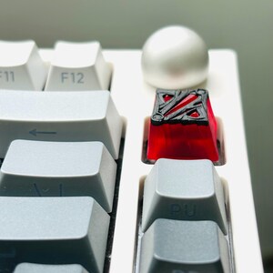 May include: A red and black keycap with a geometric design. The keycap is on a white keyboard.