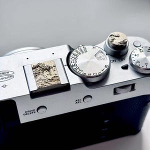 May include: A silver digital camera featuring a custom hot shoe cover and shutter button, both embossed with a textured wave design. The camera body displays silver dials with numerical markings, the text DRIVE DELETE, AEL, and a logo reading NON SYSTEM.