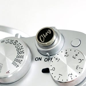 May include: Close-up of a silver camera with multiple dials and knobs. The top dial has the name "Chris" in a white script on a black background. Other dials show numbers and settings for the camera's functions.