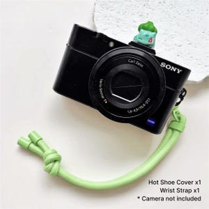 May include: Black Sony camera with a Carl Zeiss lens, a green wrist strap, and a green hot shoe cover. A small green Bulbasaur figure sits on top of the camera. The text reads: Hot Shoe Cover x1, Wrist Strap x1, * Camera not included.