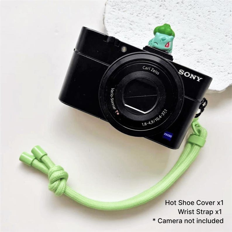 May include: Black Sony camera with a Carl Zeiss lens, a green wrist strap, and a green hot shoe cover. A small green Bulbasaur figure sits on top of the camera. The text reads: Hot Shoe Cover x1, Wrist Strap x1, * Camera not included.