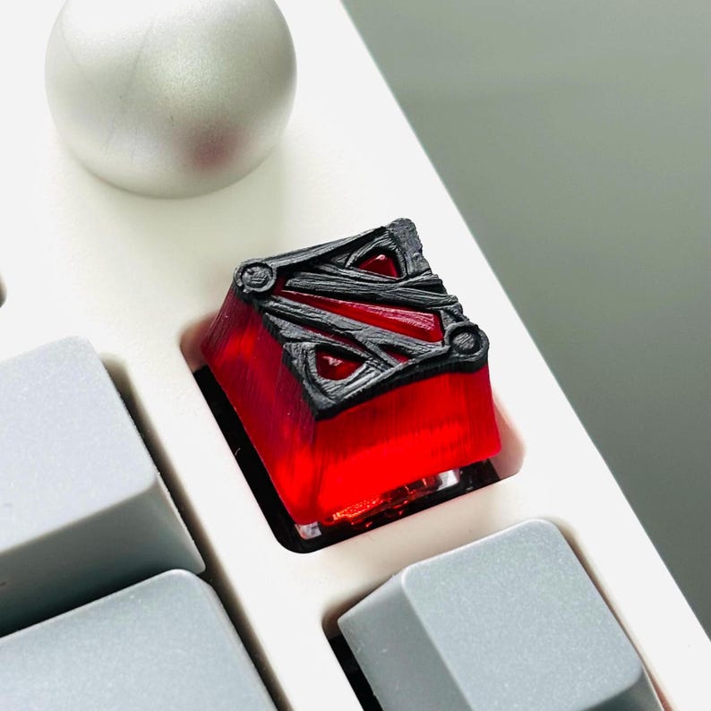 May include: A red and black keycap with a stylized logo. The keycap is made of transparent plastic and has a black logo on top.