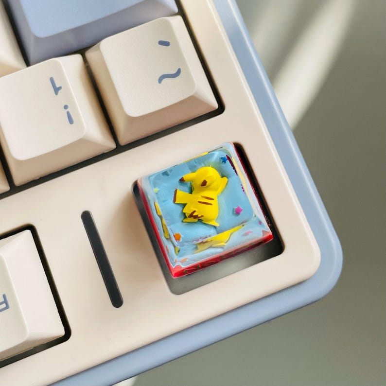 May include: A custom keyboard keycap featuring a yellow Pikachu design on a blue background. The keycap is surrounded by a red and white border, and is set on a light blue and white keyboard.