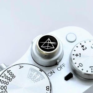 May include: A silver camera shutter button featuring a black circular inlay with a white line-art graphic of a triangular prism dispersing a beam of light. The button is mounted on a silver camera body with visible adjustment dials and text.