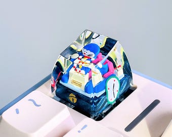 May include: A clear resin artisan keycap featuring a miniature blue and white character figure sitting at a desk with a clock graphic on the side. The keycap is mounted on a mechanical keyboard with light pink keys, showcasing a detailed design.
