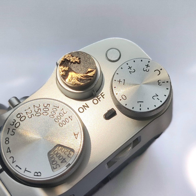 May include: A silver camera top plate featuring a circular shutter button with a gold and dark gray wave design. The camera includes a silver dial with exposure compensation markings from -3 to +3 and a shutter speed dial with numbers 2 to 5000.