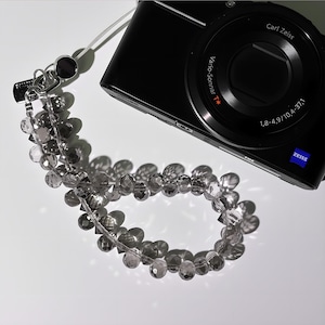 May include: A black digital camera with a silver and clear beaded camera strap. The camera has a Carl Zeiss lens and the strap is made of faceted beads. The camera strap is attached to the camera.