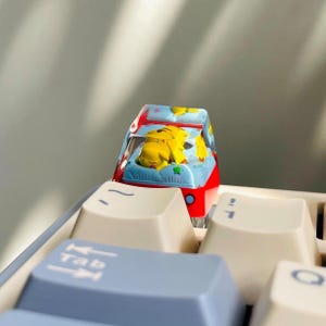 May include: A custom keyboard keycap featuring a yellow Pikachu design inside a red and blue car. The keycap is on a keyboard with white and light blue keys. The Tab key is visible.