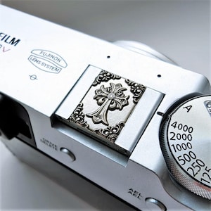 Puede incluir: Una tapa de zapata de metal plateado para cámara, con un diseño de cruz en relieve ornamentado y detalles de volutas intrincadas. El accesorio está encajado en la ranura de la zapata de un cuerpo de cámara Fujifilm de color plata.