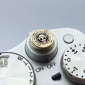 Könnte beinhalten: Ein goldfarbener Kameraknopf mit dem "One Piece"-Logo, das ein Totenkopf- und Knochen-Design in einem Kreis zeigt. Der Knopf befindet sich auf einem silbernen Kameragehäuse, mit Zifferblättern und Einstellungen.