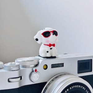 May include: A small white figurine of a puppy with black spots, wearing red-rimmed sunglasses and a red bow tie, perched on top of a silver digital camera. The puppy has pink blush on its cheeks and is positioned on the camera's hot shoe mount.