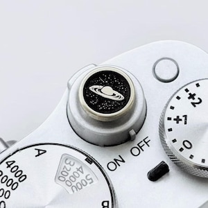 May include: A silver camera shutter button featuring a circular black inlay with a white graphic of the planet Saturn surrounded by stars. The button is mounted on a silver camera top plate with the text ON OFF and a silver exposure dial nearby.